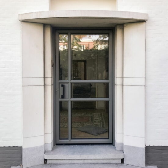 Reynaers SL38 Aluminium Voordeur – Modern aluminium Steel Look voordeur met Strakke Asymmetrie en Elegant Lijnenspel, Grijze Deur Tegen Lichte Bakstenen Gevel.