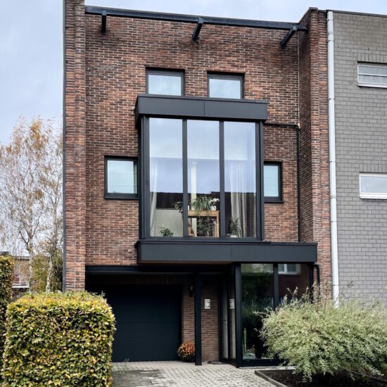 Voorgevel woning met een moderne zwarte aluminium erker met een vlakke dakrand op eerste verdiep. Moderne zwarte aluminium ramen en deuren met een aluminium en ceder oversteek en garagepoort.