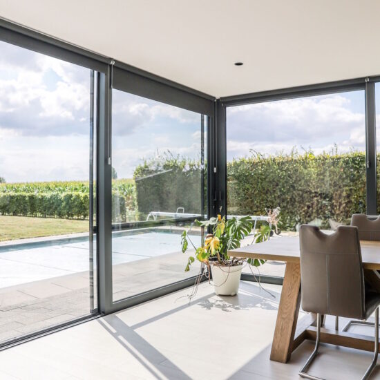 De woonveranda beschikt over grote schuifdeuren en schuiframen, waardoor er een optimale lichtinval ontstaat en de tuin naadloos wordt verbonden met de leefruimte. Het hoge plafond geeft de veranda een ruim en luchtig gevoel, waarbij het minimalistische en moderne ontwerp wordt benadrukt door het gebruik van zwarte aluminium Reynaers profielen. De strakke lijnen en het sobere ontwerp zorgen voor een stijlvolle uitstraling, terwijl het grote glas optimaal uitzicht biedt op de zonnige tuin. De combinatie van comfort, functionaliteit en esthetiek maakt deze veranda een prachtige uitbreiding van de woonruimte, perfect voor genieten van het buitenleven binnen een moderne en lichte omgeving.
