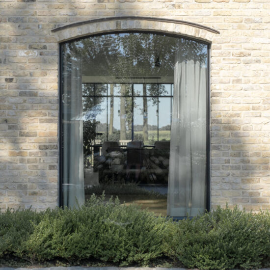 Een aluminium boograam in een klassieke gevel van rustieke bakstenen, omgeven door natuur. Het raam heeft een elegant profiel dat naadloos past bij de historische uitstraling, aangevuld met lichte hanggordijnen voor een zachte sfeer. De natuurlijke omgeving benadrukt de rustieke charme van de gevel.