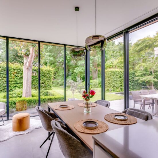 Binnenzicht van een zwarte aluminium veranda dat dient als de eetruimte naast de keuken. Een houten eettafel met zwarte aluminium tafelpoten en afgeronde stoffen vintage zetels als blikvanger. Versiert met rode bloemen in een glazen potje met water, 4 ronde rieten eetmatten met een bordje uit keramiek bovenop. De tafel is verlicht met 2 vintage stijl ronde glazen lampen. Op de achtergrond de aluminium raam en vouwwand met dubbel glas van de veranda, met uitzicht op de tuin met kort gemaaid lichtgroen gras, omringd door struiken en bomen.